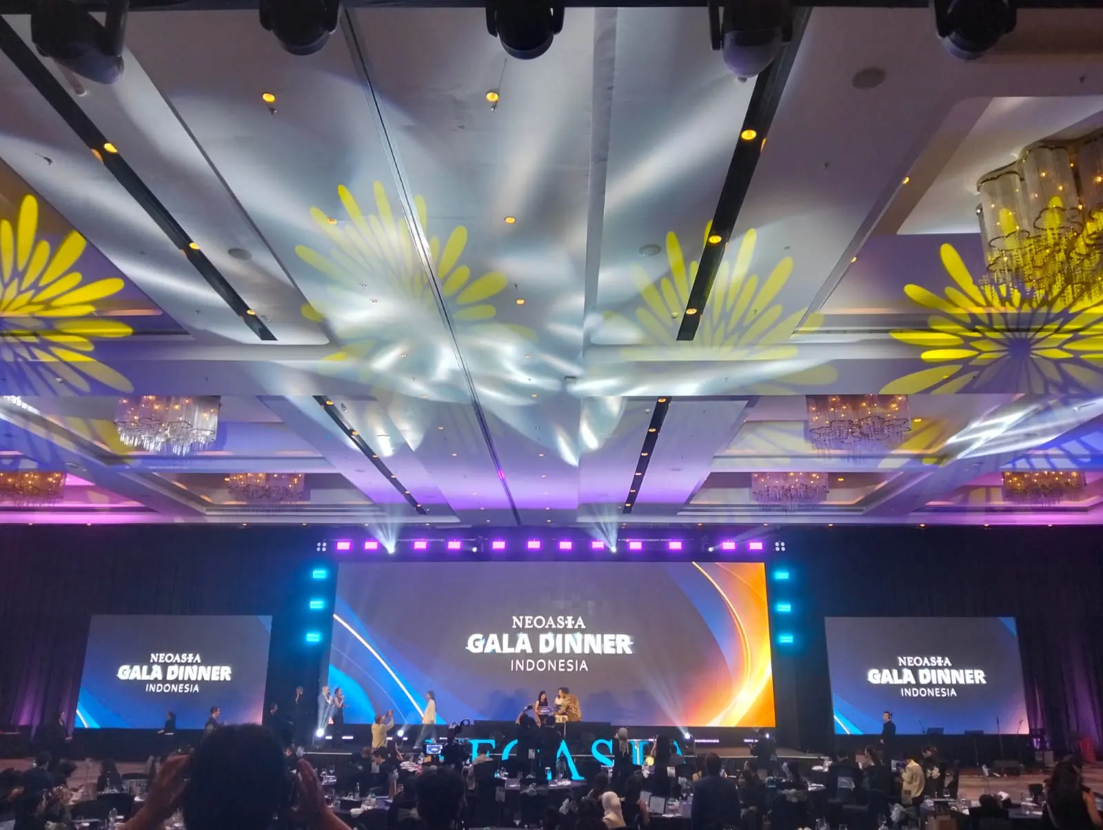 Panggung Neoasia Gala Dinner dengan tiga layar dan cahaya kuning.