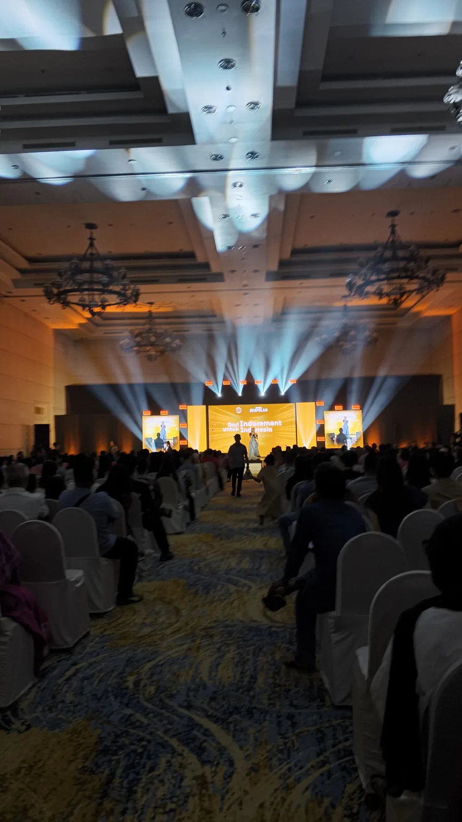 Layar LED dan sorot lampu beam di panggung acara Indocement di Cirebon, dengan penonton di ballroom.